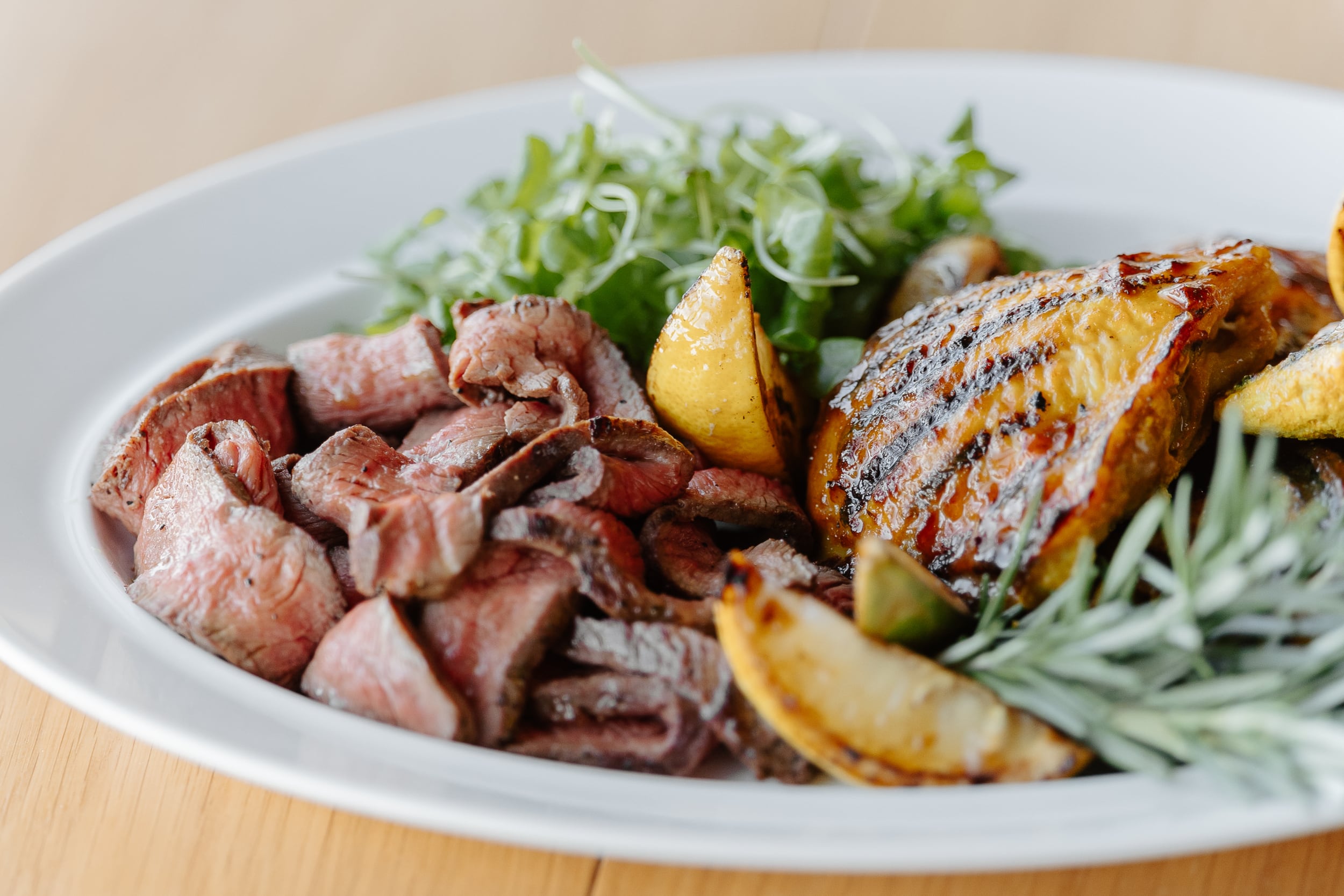 A white plate with sliced medium-rare beef, grilled chicken, roasted potato wedges, fresh greens, and rosemary garnish. The dish is set on a wooden table.