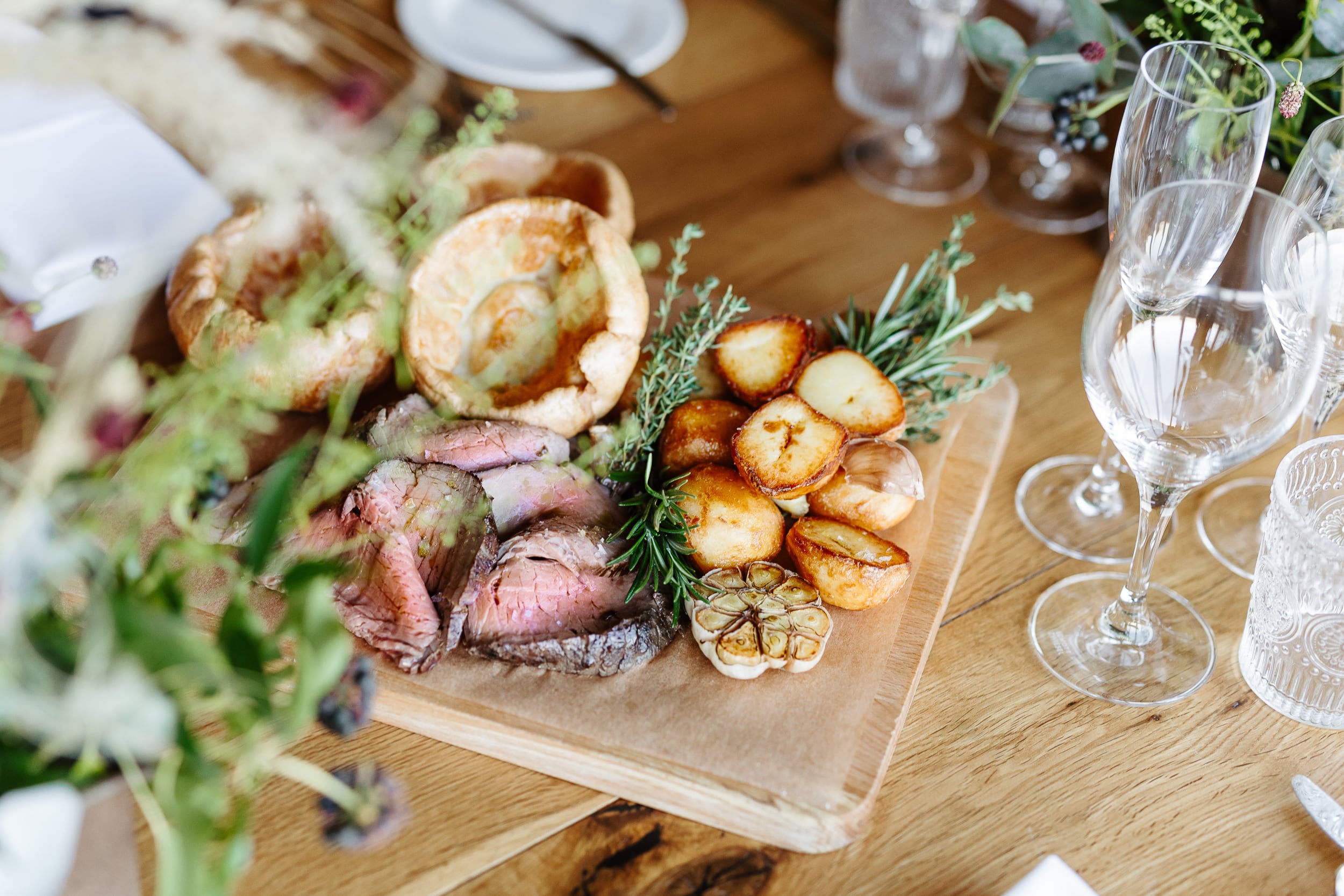 A wooden board on a table holds slices of roast beef, roasted potatoes, garlic, and Yorkshire puddings, garnished with rosemary. Nearby are empty champagne flutes and other glassware, with floral decorations partially visible.